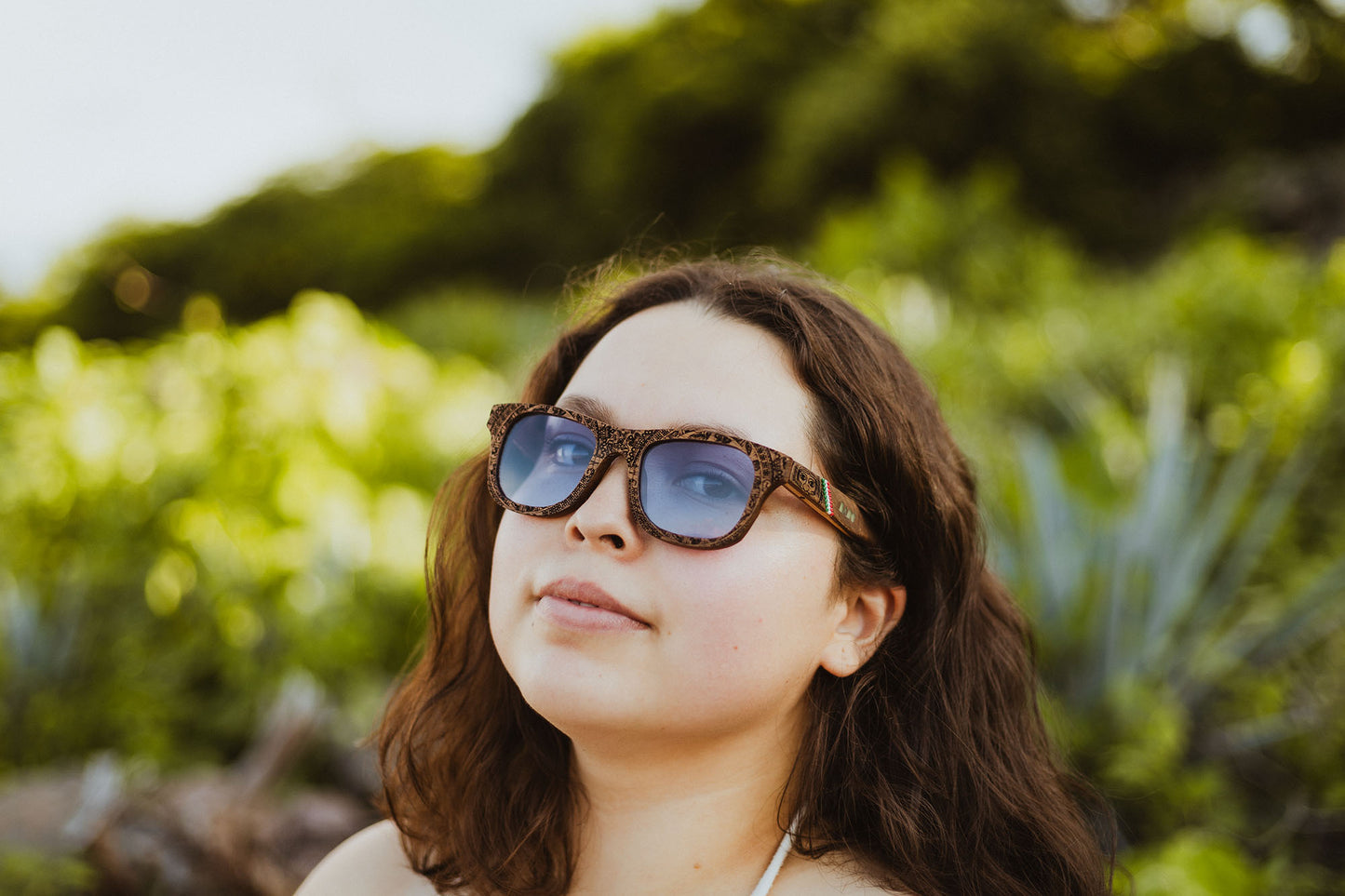 Modelo mujer usando lentes de madera Bang, modelo Mexa, con el calendario azteca grabado, intervenidos con artesanía chaquira huichol, mica morada, fondo de naturaleza