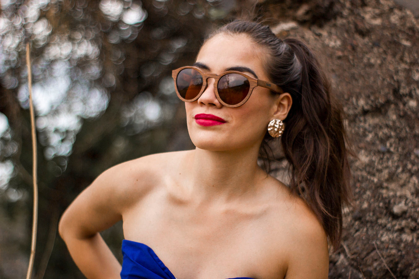 Modelo mujer usando lentes polarizados de madera, marca bang, mica café polarizada, fondo de naturaleza con bokeh