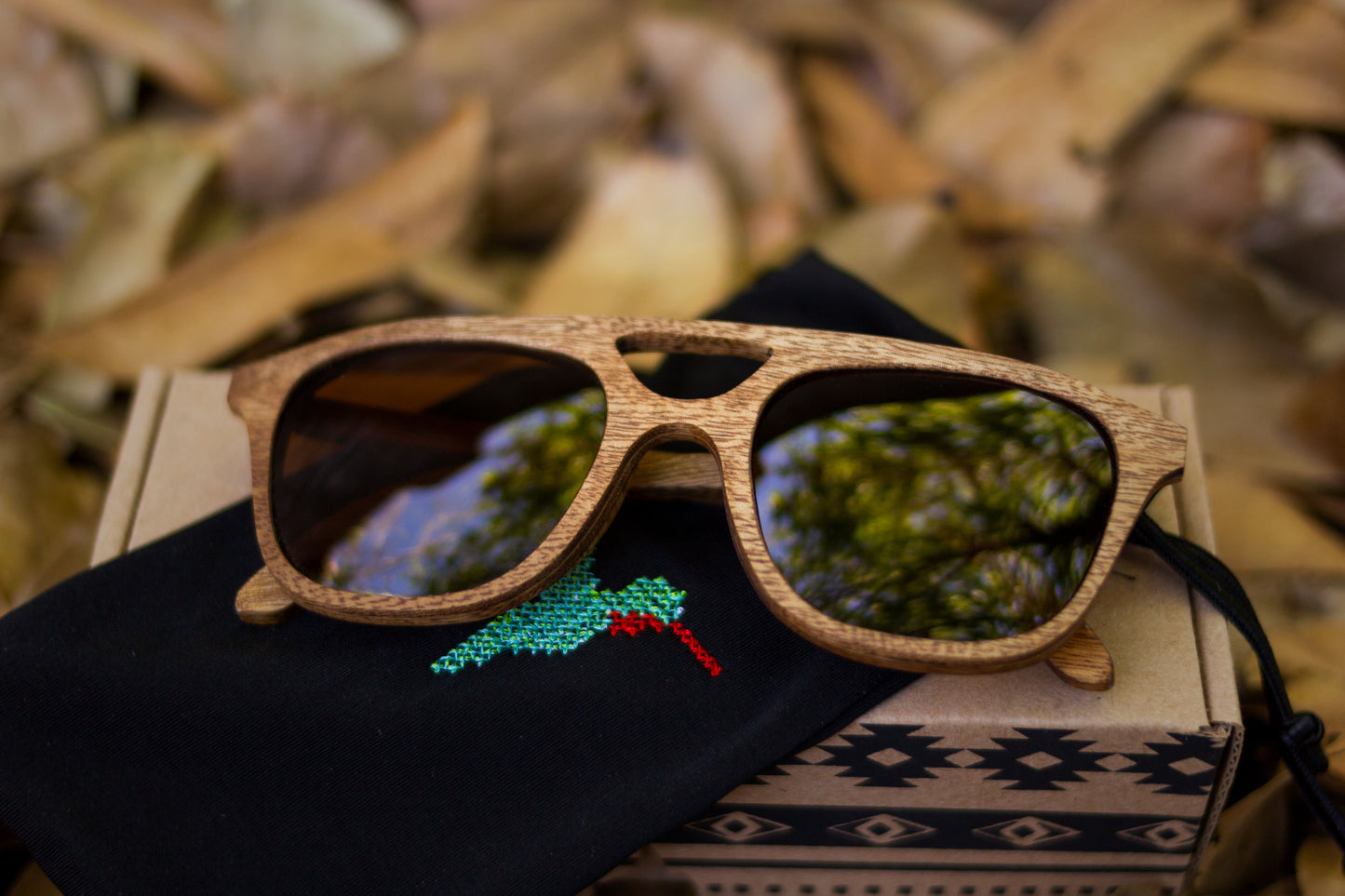 Lentes de madera bang, modelo cobá, madera rosa morada, mica café polarizada, sobre su empaque de cartón y su morral negro bordado con un colibrí
