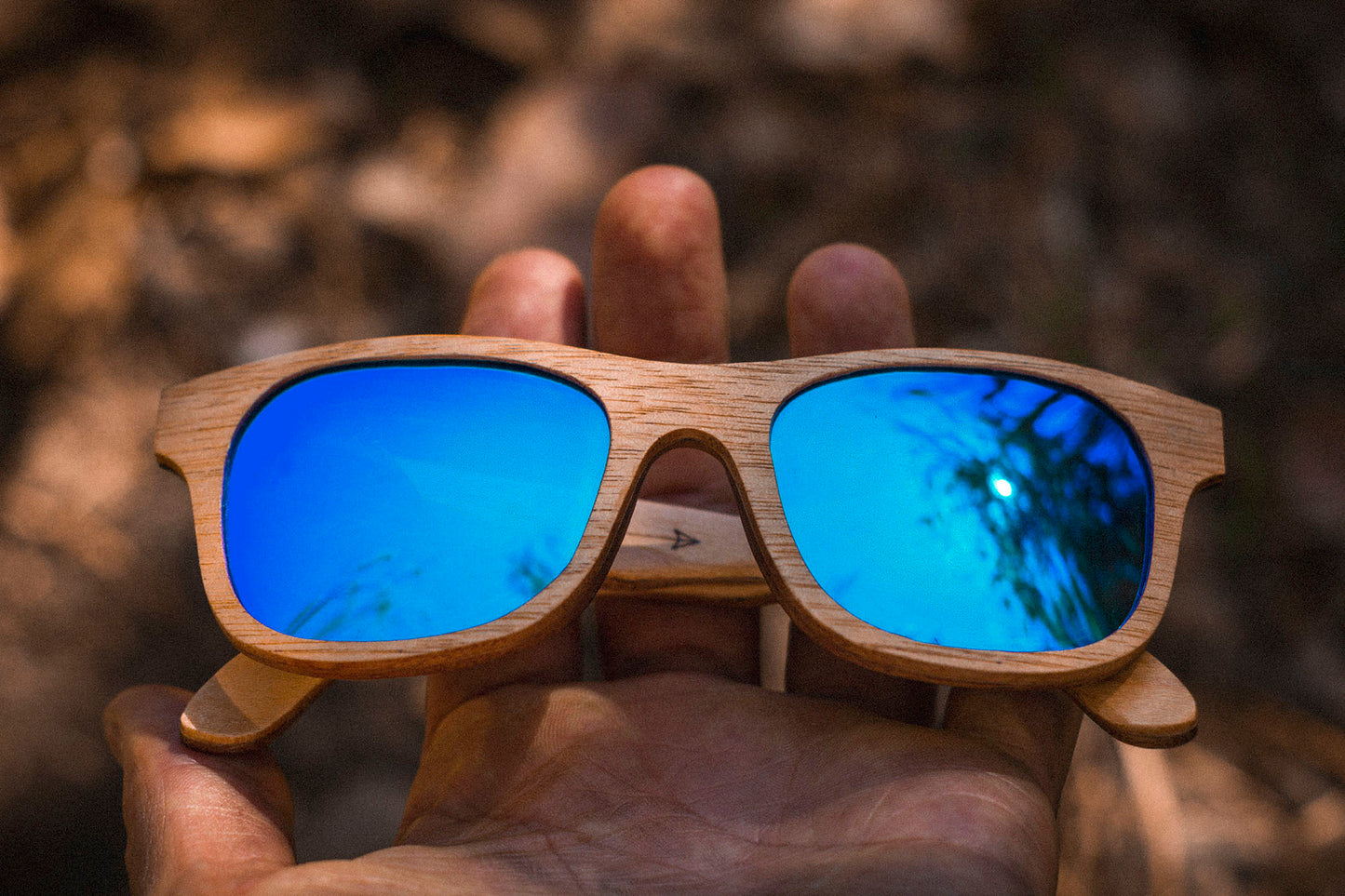 Manos sosteniendo unos Lentes de madera bang, micas polarizadas azul espejo, modelo nativo, fondo café