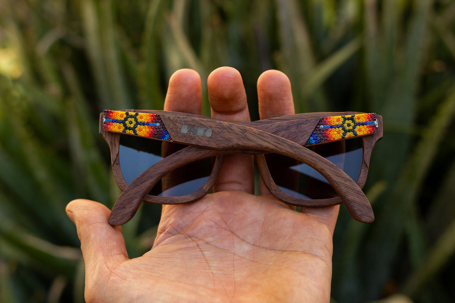Mano sosteniendo unos lentes wirra, modelo hikuri sol, con artesanía huichol, mica oscura, fondo de naturaleza con bokeh