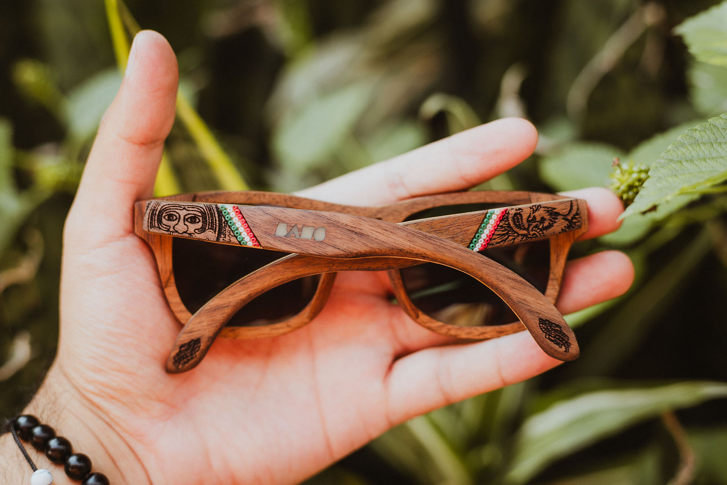 Mano sosteniendo unos lentes de madera conmemorativos de México, intervenido con artesanía huichol, fondo de naturaleza 