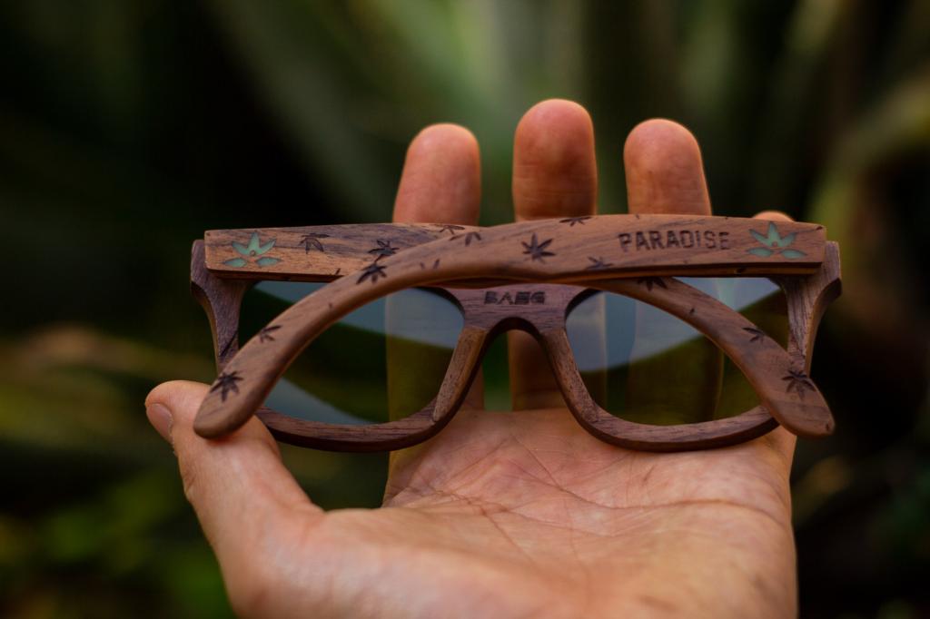 Mano sosteniendo lentes paradise de bang, edición especial, grabado de cannabis, logo que brilla, hechos de madera, fondo de vegetación bokeh